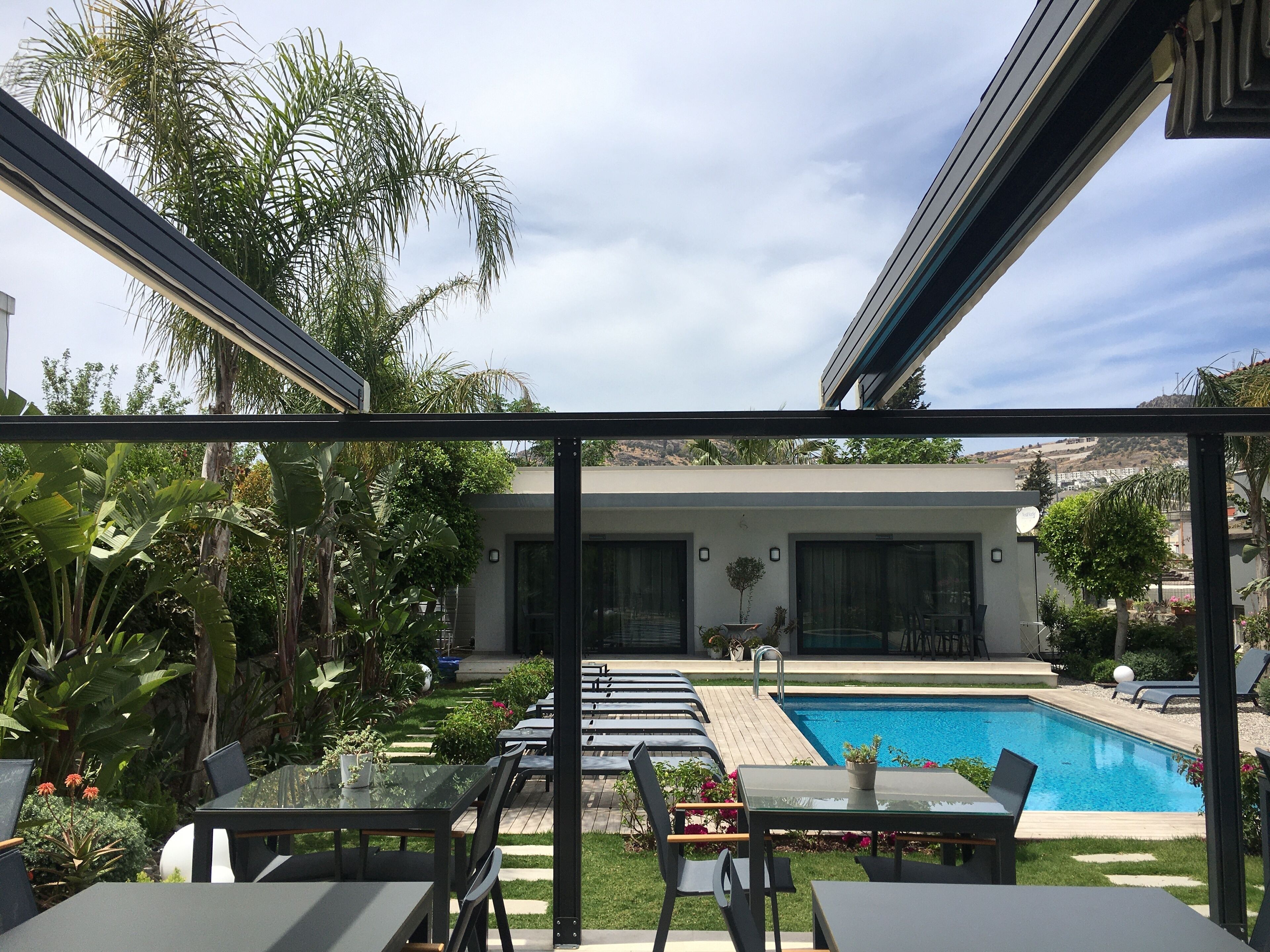 A modern outdoor pool area with lounge chairs and tables, surrounded by lush greenery and a clear blue sky.
