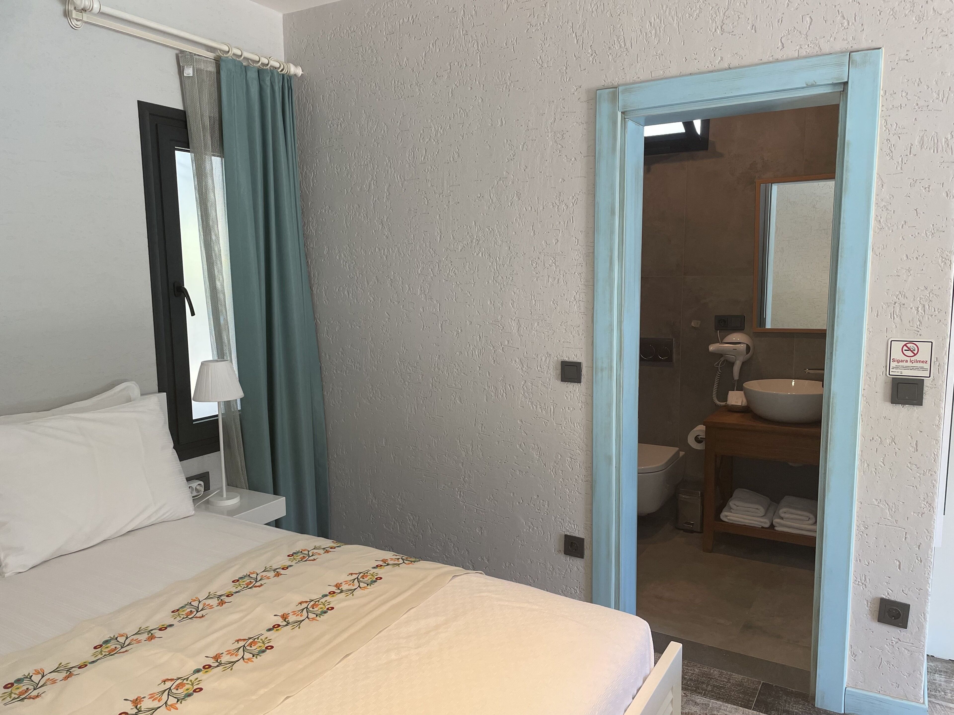 Une chambre d’hôtel avec un lit, une fenêtre avec des rideaux, une salle de bain visible à travers une porte ouverte, et un panneau indiquant qu’il est interdit de fumer.