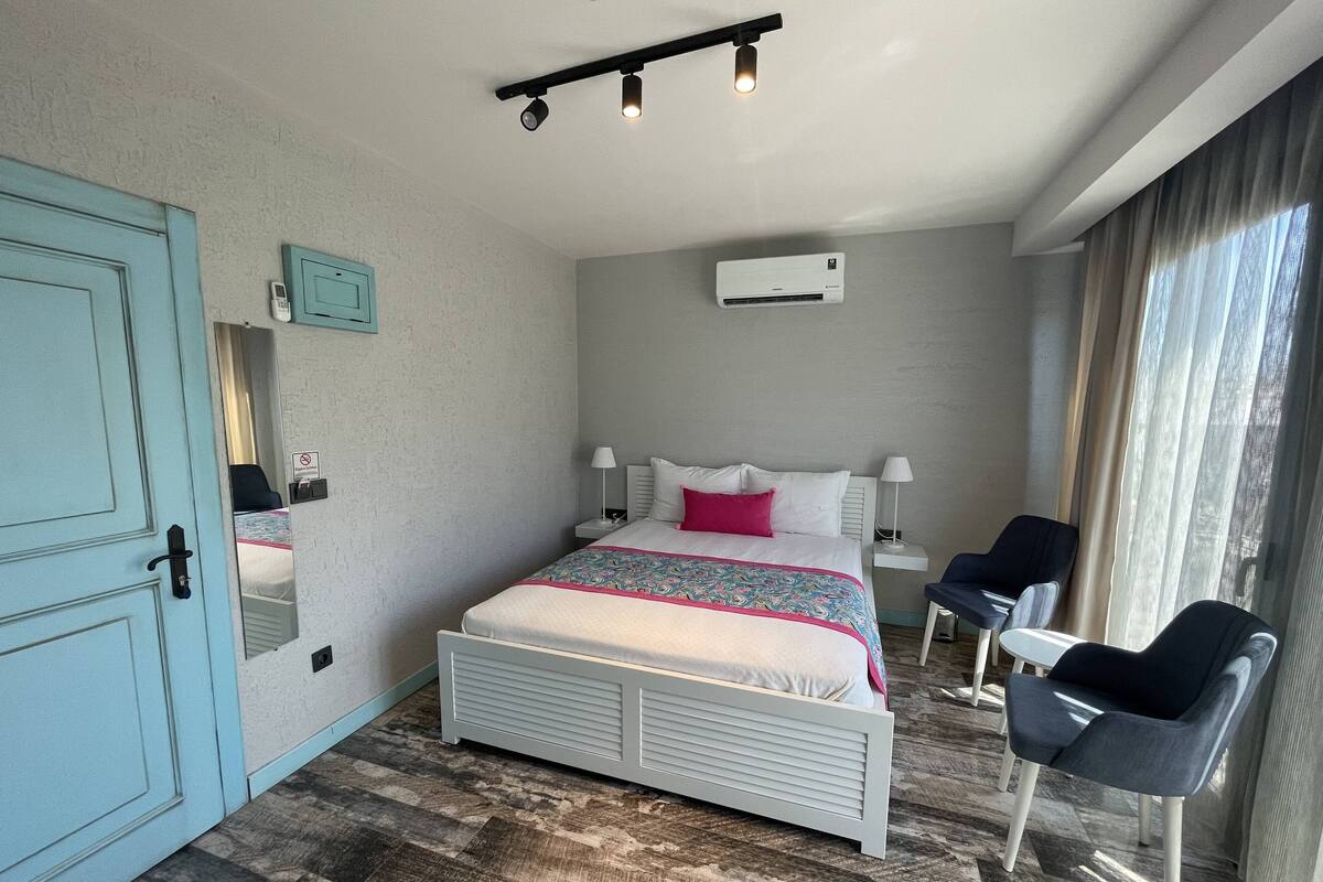 Une chambre à coucher comprenant un lit, des tables de chevet, une chaise, une fenêtre avec des rideaux, un appareil de climatisation et une porte.