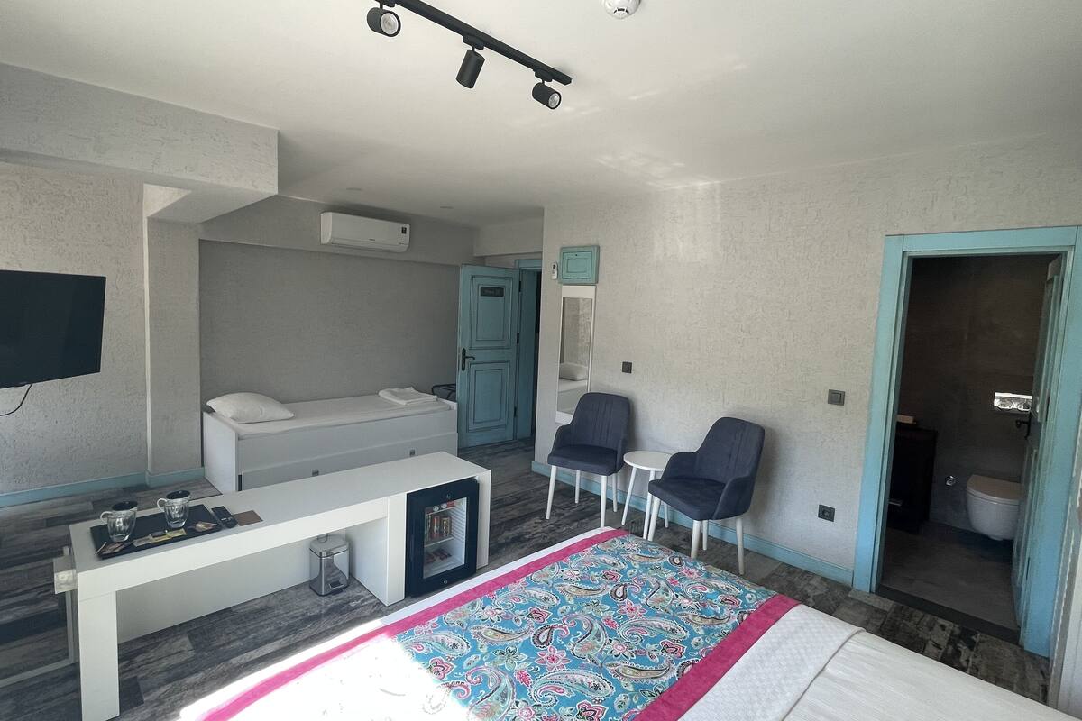 Une chambre d’hôtel moderne équipée d’un lit, d’une télévision, d’un bureau, de deux chaises, d’une petite table et d’une salle de bain visible à travers une porte ouverte.