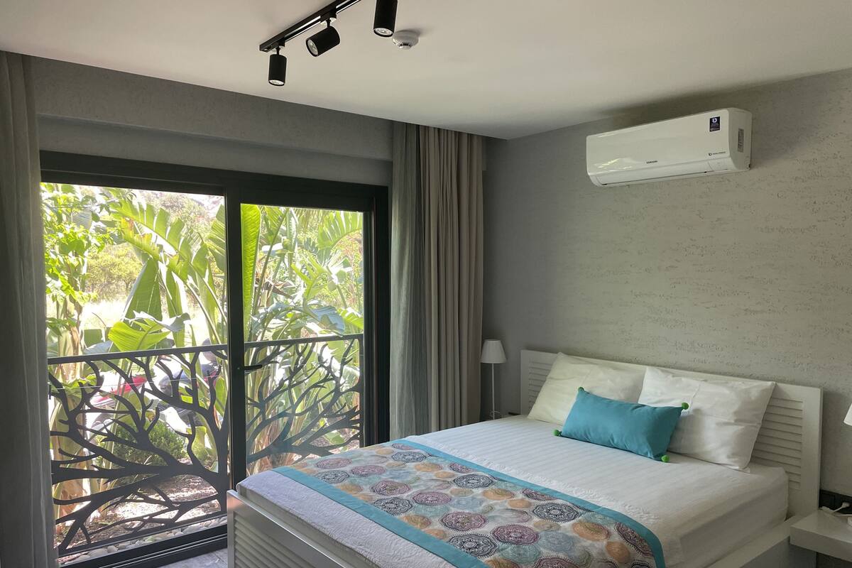 Une chambre d’hôtel avec un lit, une fenêtre donnant sur un espace verdoyant, un appareil de climatisation et un luminaire au plafond.