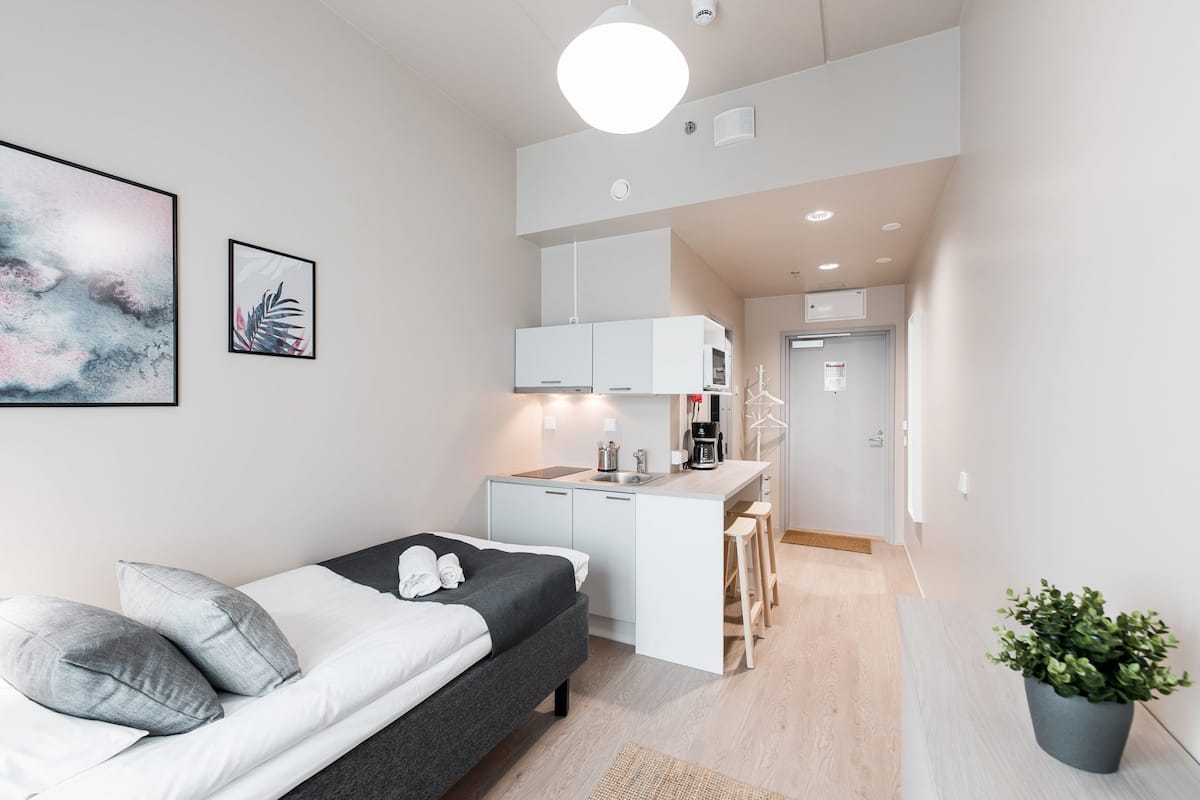 A modern hotel room with a bed, a small kitchenette, a desk, and a potted plant.