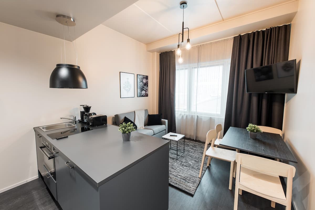 A modern kitchen with a central island, a dining area with a table and chairs, a flat-screen TV mounted on the wall, and a large window with curtains.