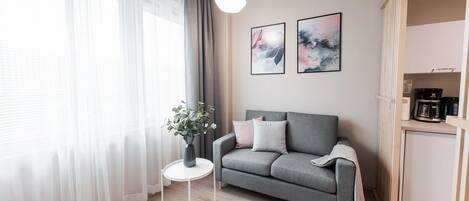 Moderni olohuone, jossa on harmaa sohva, pieni valkoinen sivupöytä maljakolla ja suuri ikkuna, jossa on läpikuultavat verhot.