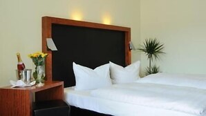 Une chambre d’hôtel avec un grand lit, une tête de lit en bois, une table de chevet avec une bouteille de champagne et des fleurs, et une plante en pot.