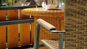 Une chaise et une table en osier, avec une bouteille et des verres, sont placées sur un balcon donnant sur une colline verdoyante parsemée de maisons.