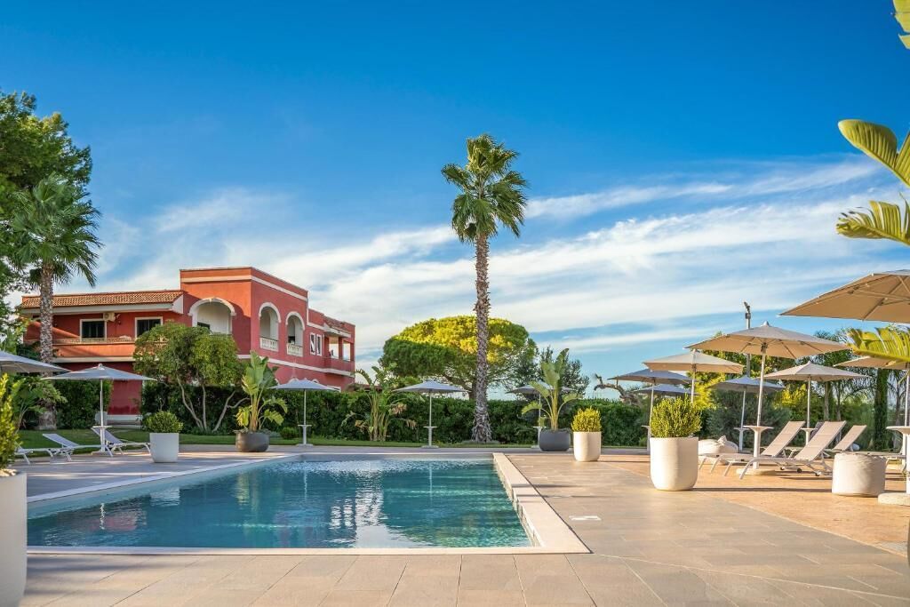 Una piscina con sillas de descanso y sombrillas, rodeada de palmeras y un edificio rojo.