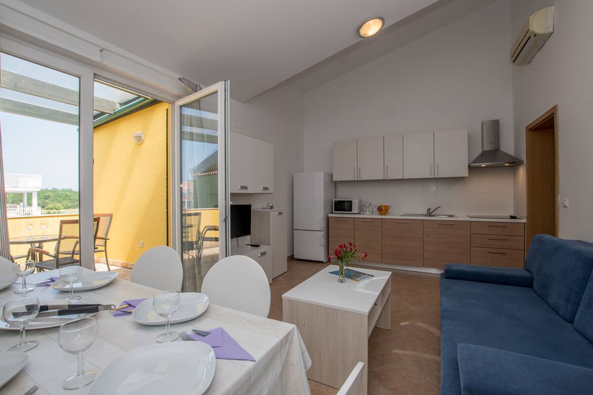 Cozinha moderna com armários brancos, micro-ondas e pia. Há uma área de jantar com mesa posta para refeição, e um sofá azul na sala.