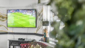 Huoneessa on suuri, seinälle asennettu litteänäyttöinen televisio, jossa näkyy jalkapallo-ottelu, nahkasohva, pieni pöytä ja koristeellinen seinäkoriste.