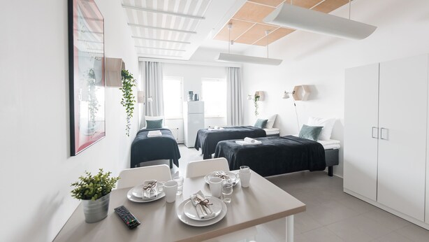 Et moderne hotelværelse med spiseplads, to senge, et garderobeskab og et loft med træpaneldesign.