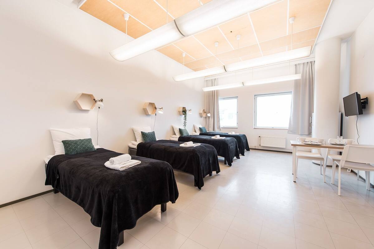 A hotel room with four beds, each with a black bedspread and white towels, a desk with a chair, and a large window providing natural light.