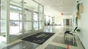 En moderne hotellobby med glasdøre, en sort dørmåtte, et siddeområde og en plante på væggen.