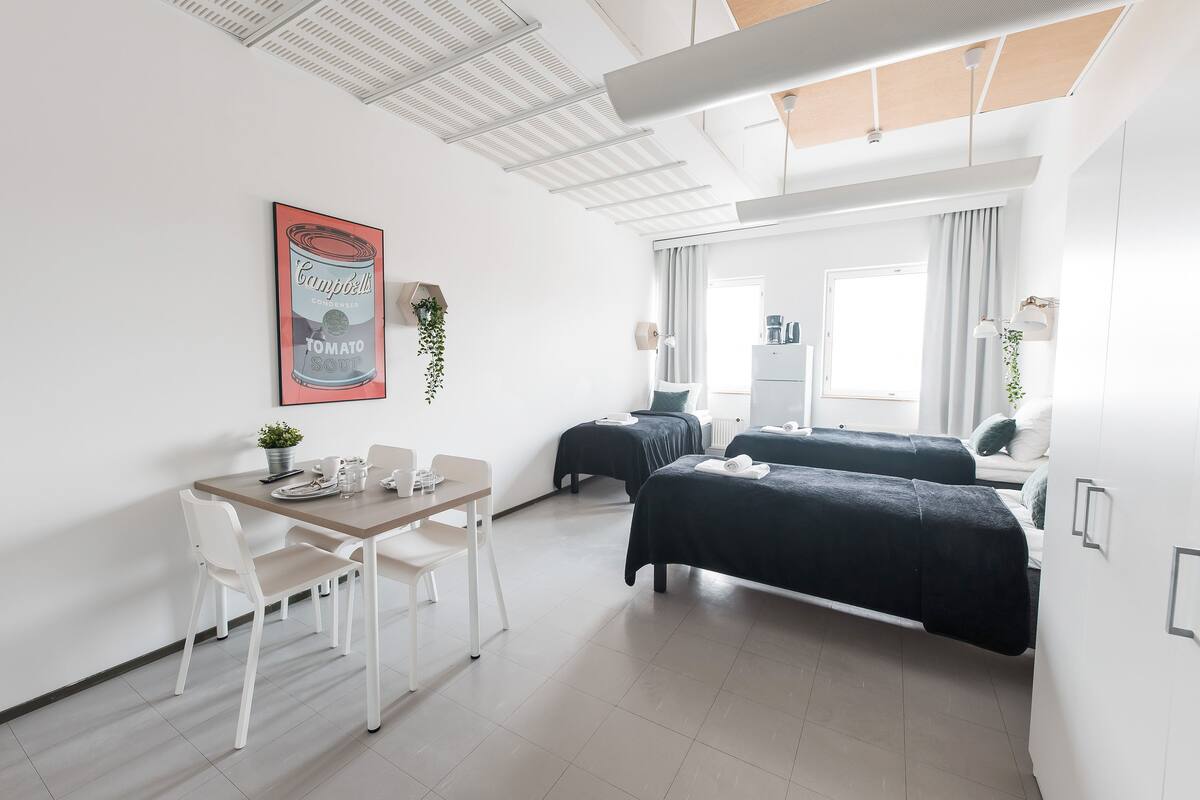 A modern hotel room with a dining table, two beds, a ceiling light, and a window with curtains.