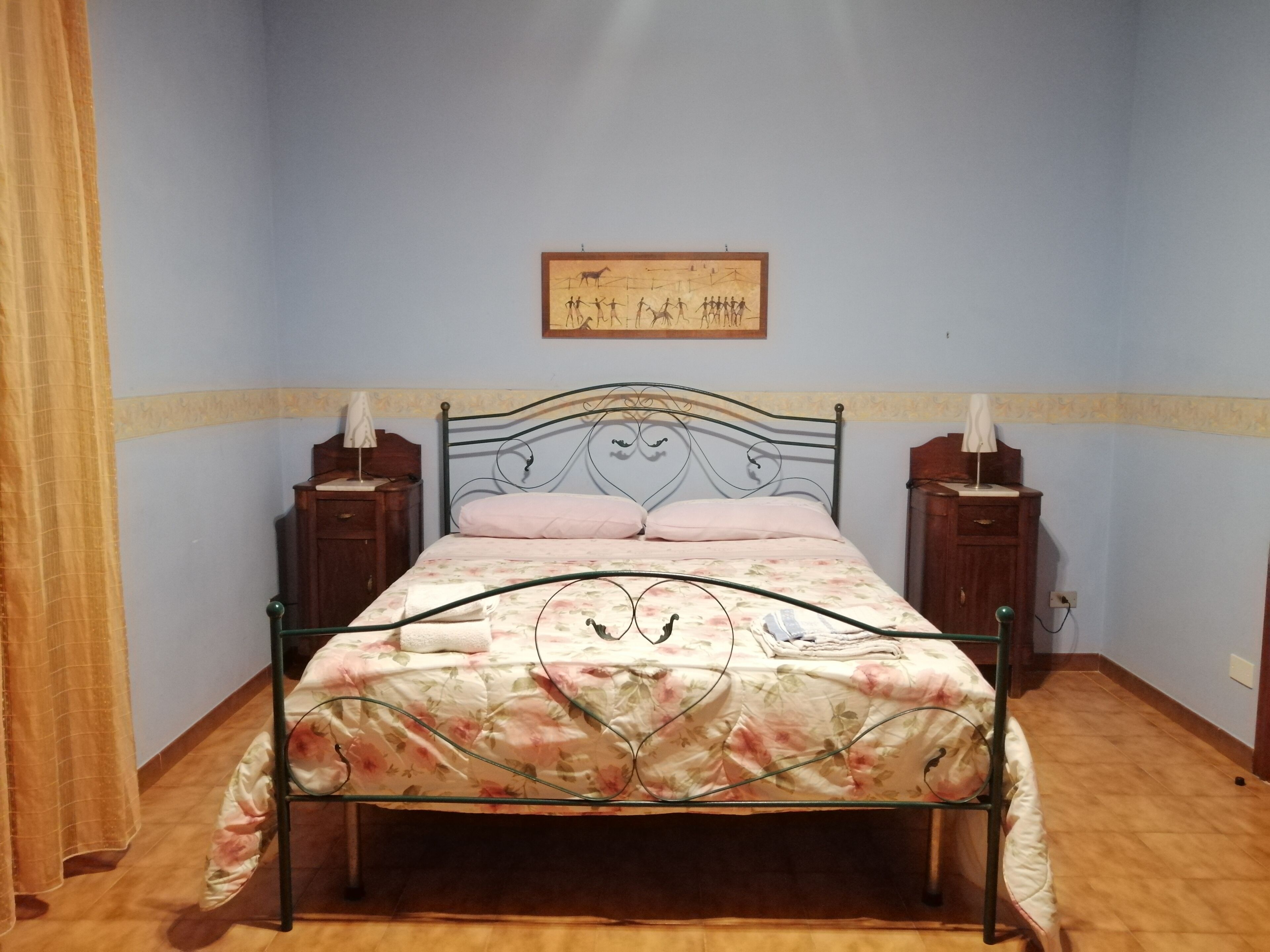 A bedroom with a metal-framed bed, two bedside tables with lamps, a wooden headboard, and a framed picture on the wall.