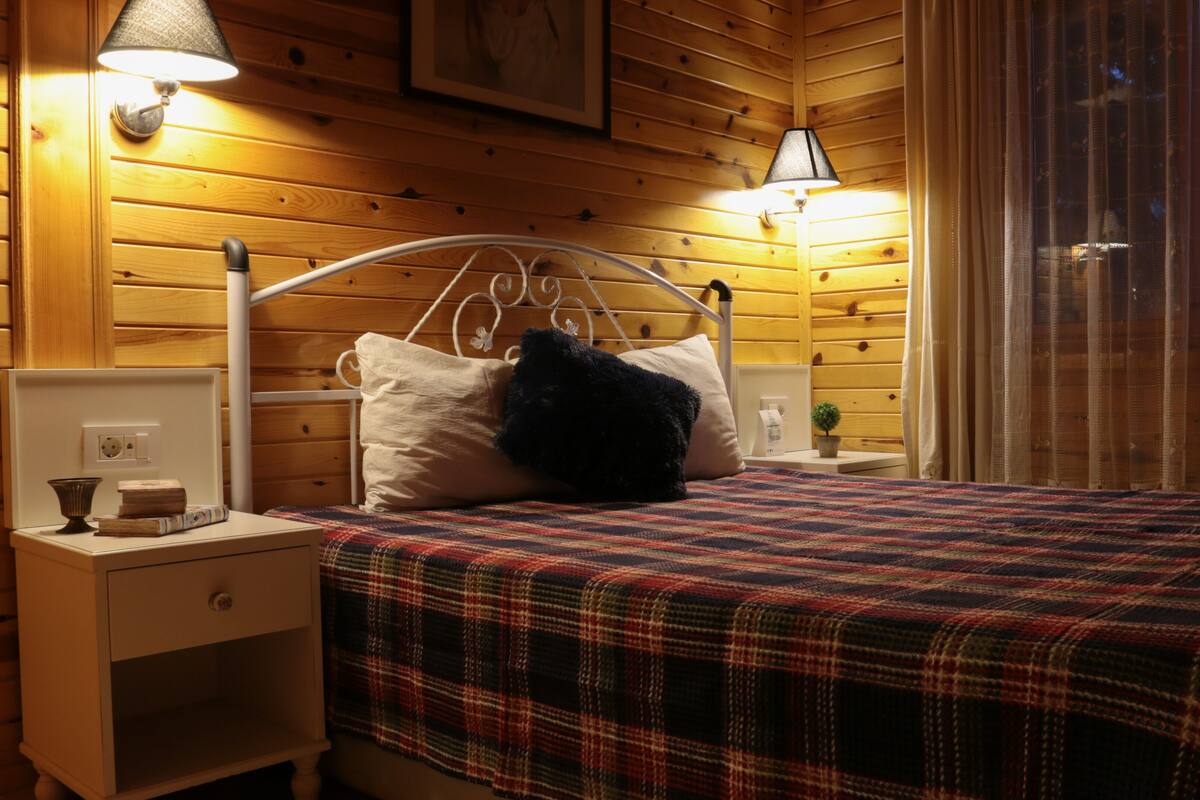 A wooden cabin bedroom with a bed, two bedside lamps, a nightstand, and a small table with a lamp.