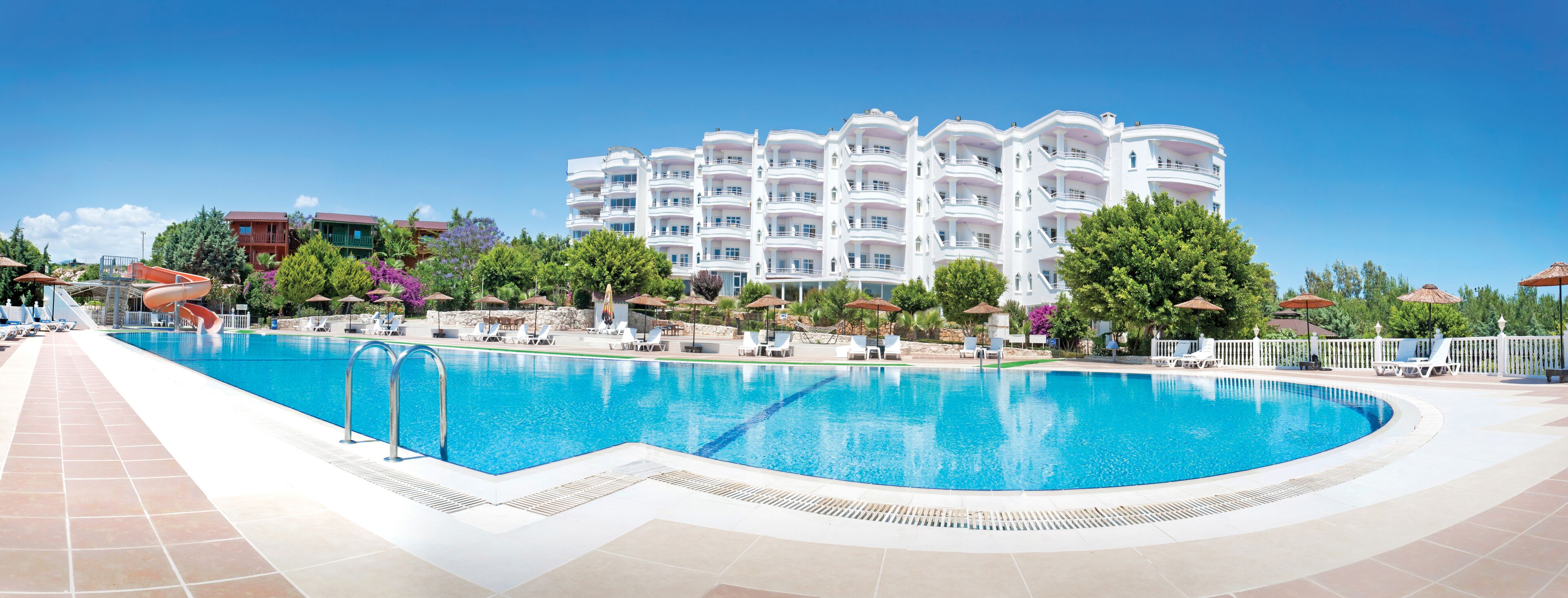 A large swimming pool with lounge chairs and umbrellas, surrounded by a white building with balconies.