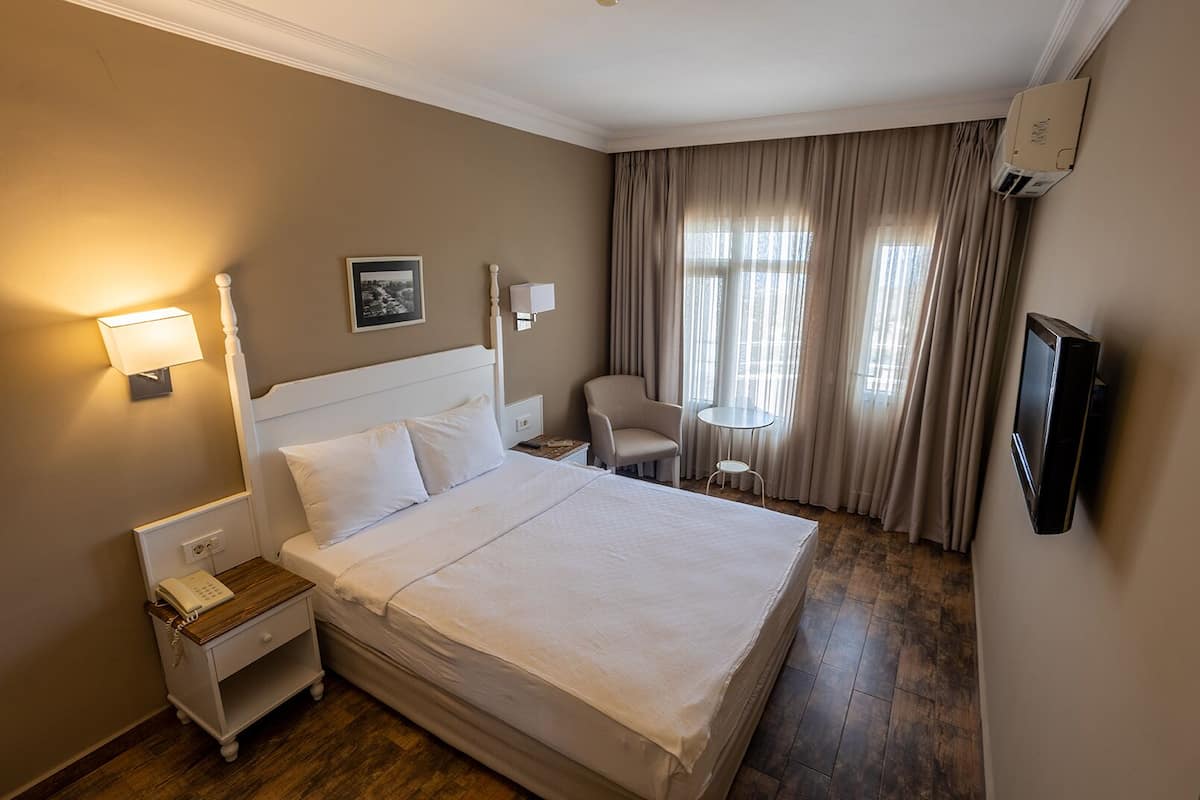 A hotel room with a large bed, bedside tables, a chair, a small table, a TV, and a window with curtains.
