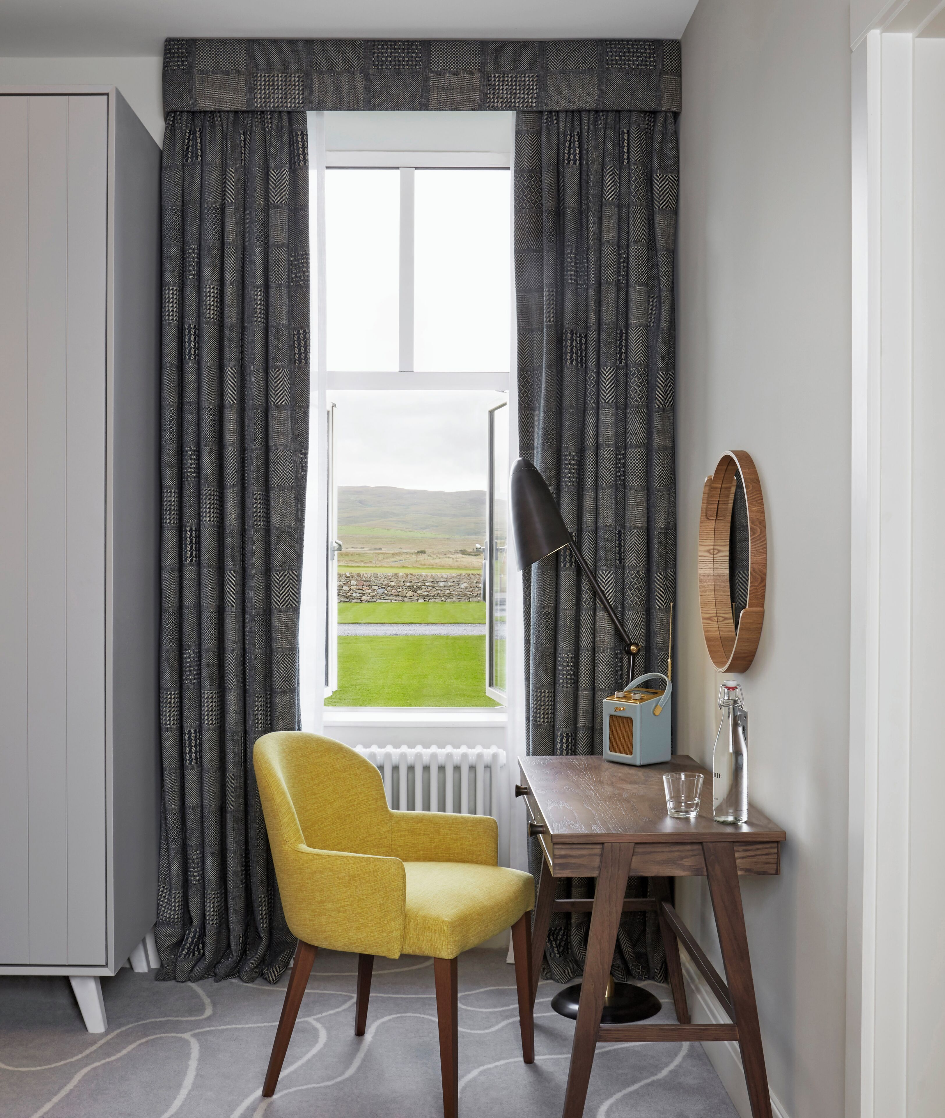 A room with a view of a green field through a window, a yellow chair, a wooden desk, a lamp, a radio, and a bottle.