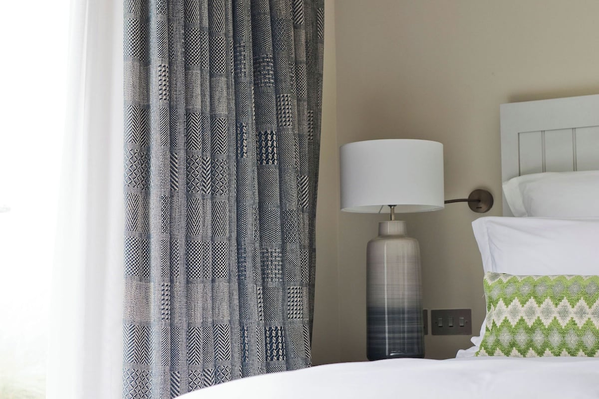 A neatly made bed with white bedding, a lamp on a bedside table, and patterned curtains.