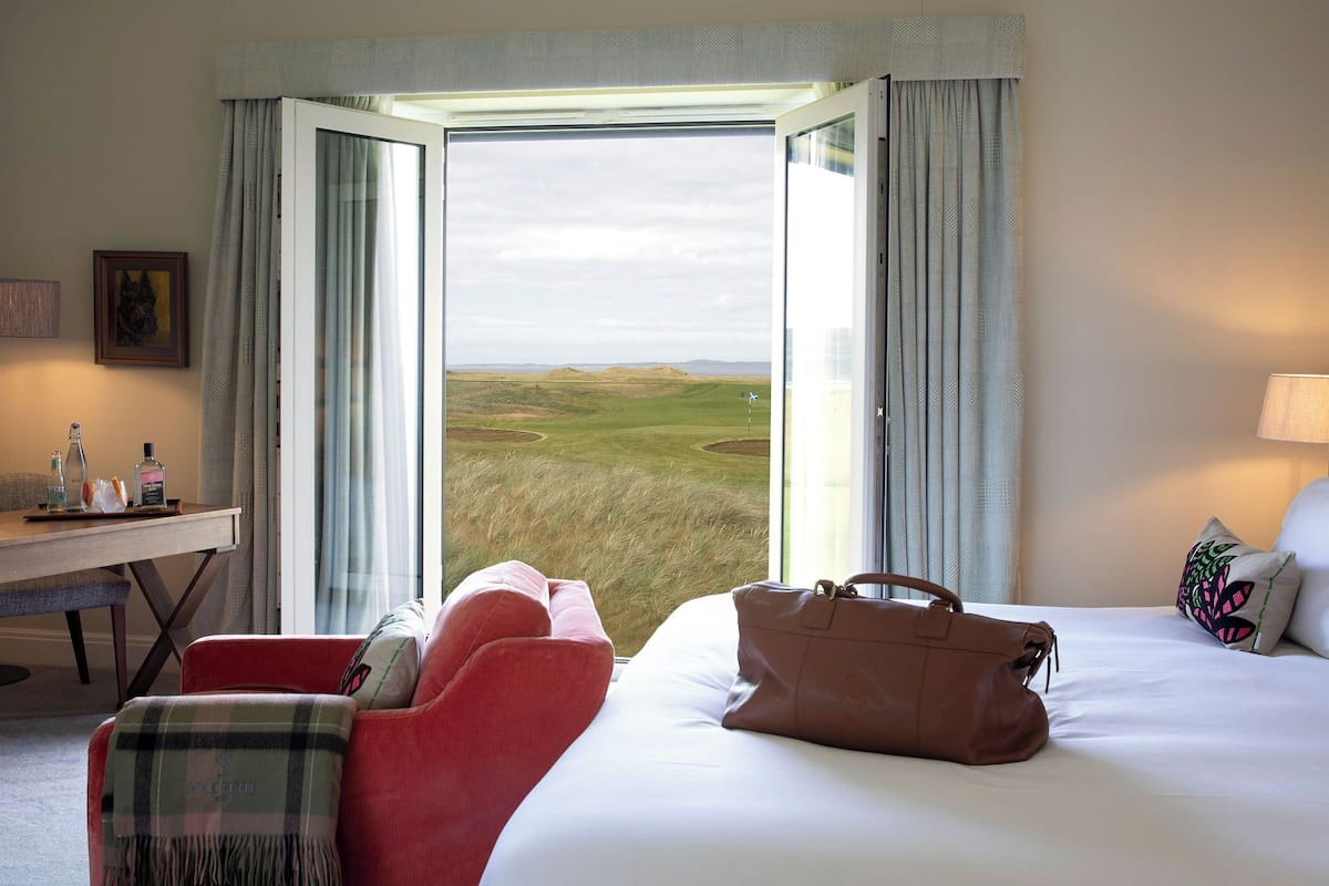 A hotel room with a bed, a red chair, a brown handbag, a desk with a lamp, and a view of a grassy landscape through an open window.