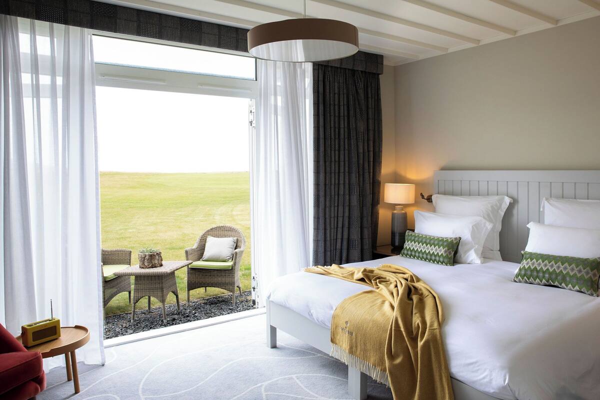 A hotel room with a bed, a nightstand, a lamp, a chair, a small table, and a view of the outdoors.