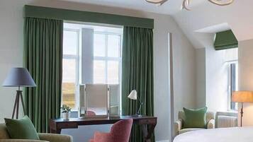 Habitación de hotel con una cama grande, un escritorio con una silla, una mesita pequeña y otra silla. Se aprecia una ventana con cortinas, un candelabro y una lámpara sobre el escritorio.