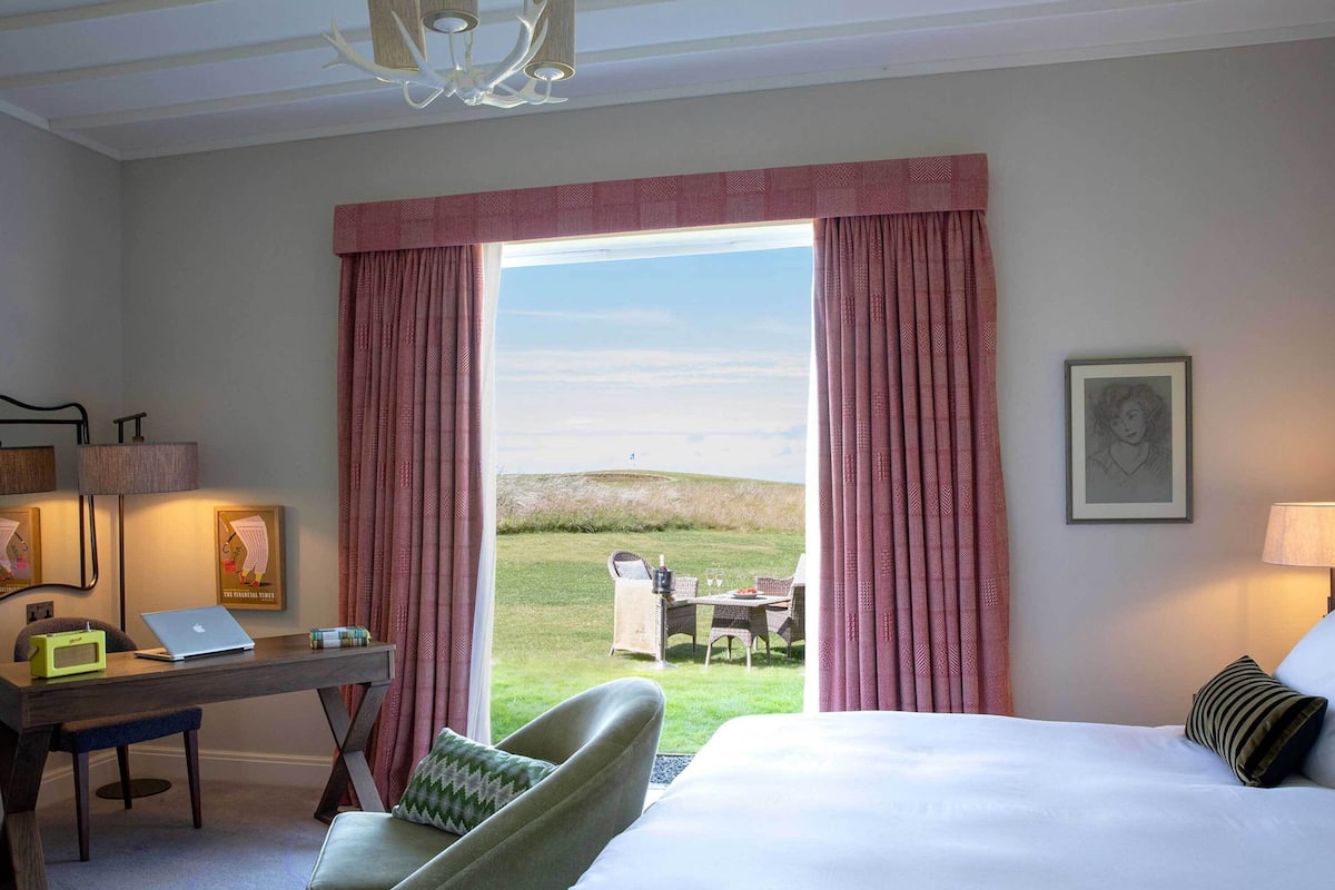 A hotel room with a bed, a desk with a laptop, a chair, and a view of a grassy area through a window.