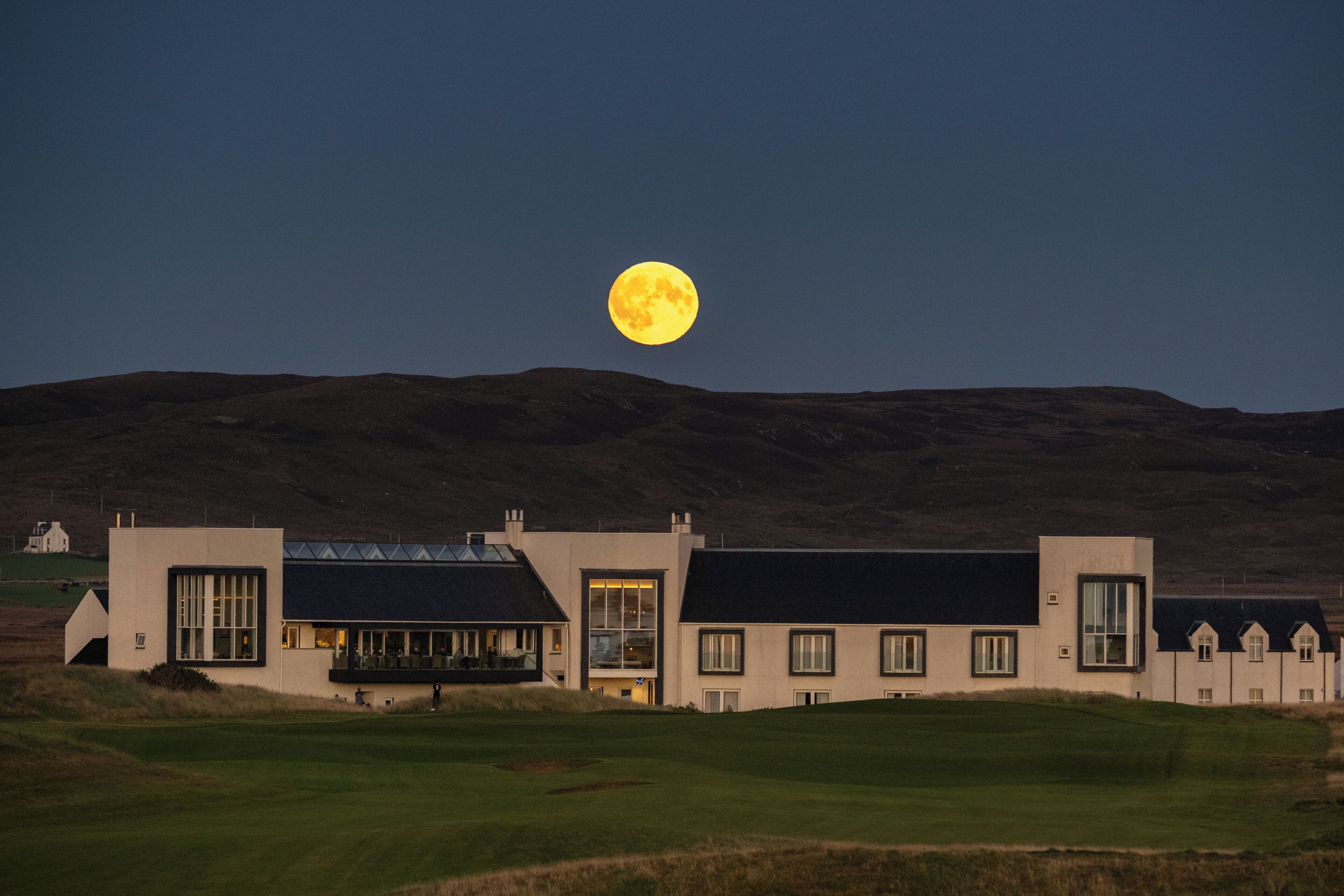 A building with a flat roof and large windows, set against a backdrop of hills and a full moon.