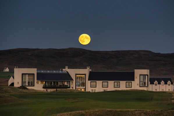 A building with a flat roof and large windows, set against a backdrop of hills and a full moon. - Another Place, The Machrie (Islay Island)