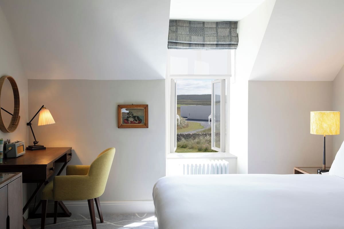 A hotel room with a bed, a desk with a chair, a lamp, a picture on the wall, and a window with a view of the countryside.