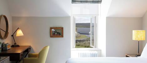 Habitación de hotel con cama, escritorio y silla, lámpara, un cuadro en la pared y ventana con vistas al campo.