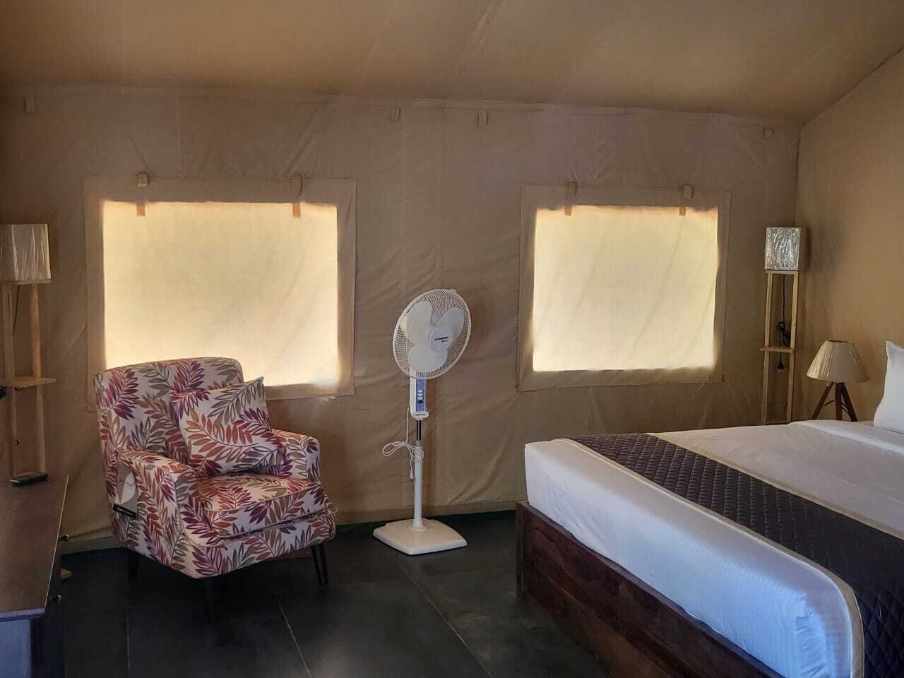 A room with a bed, a chair, a standing fan, and two windows with curtains.