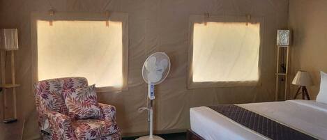 A room with a bed, a chair, a standing fan, and two windows with curtains.