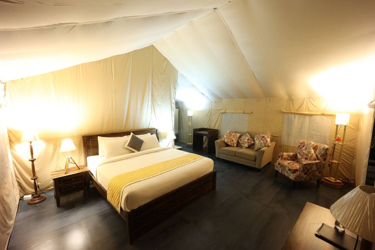 Habitación tipo alojamiento en tienda de campaña, con cama, mesita de noche, lámpara, silla y una pequeña mesa con lámpara.
