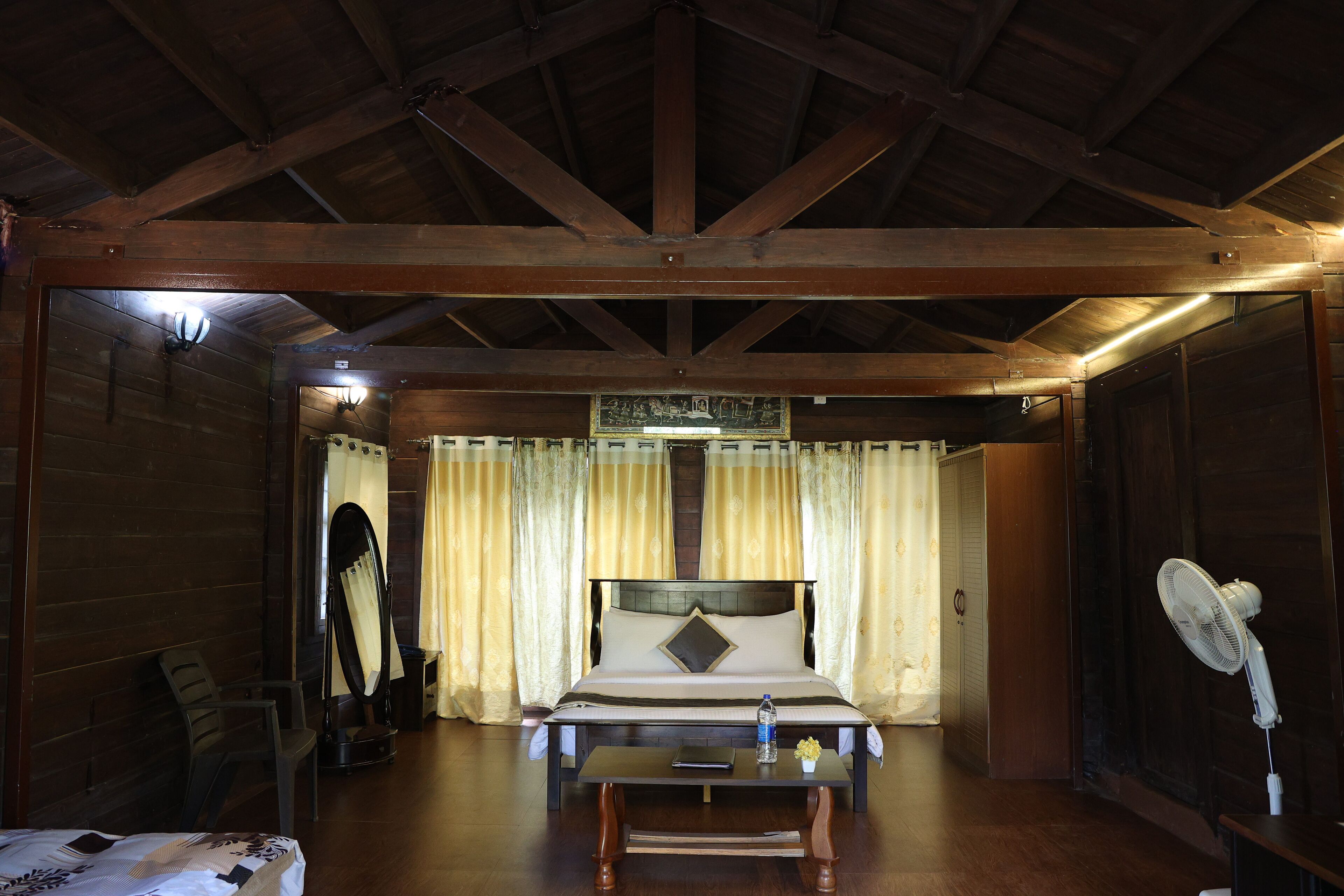 A wooden-framed room with a bed, a table, a fan, and curtains.