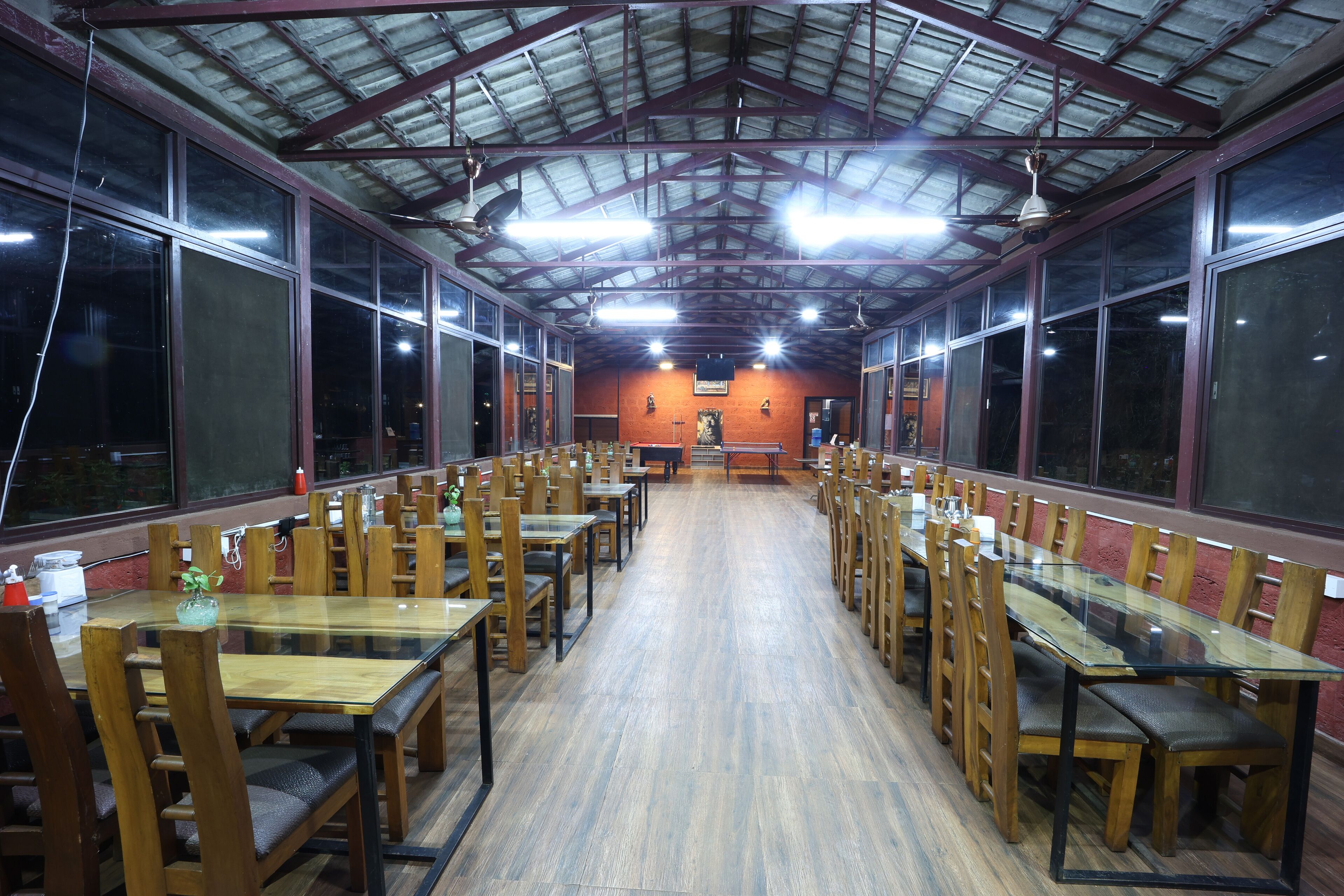 A spacious dining area with long wooden tables and chairs, large windows, and a high ceiling with metal beams.