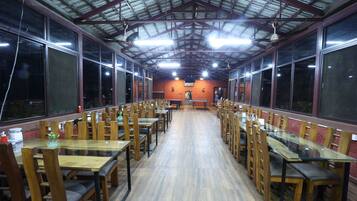 A spacious dining area with long wooden tables and chairs, large windows, and a high ceiling with metal beams.