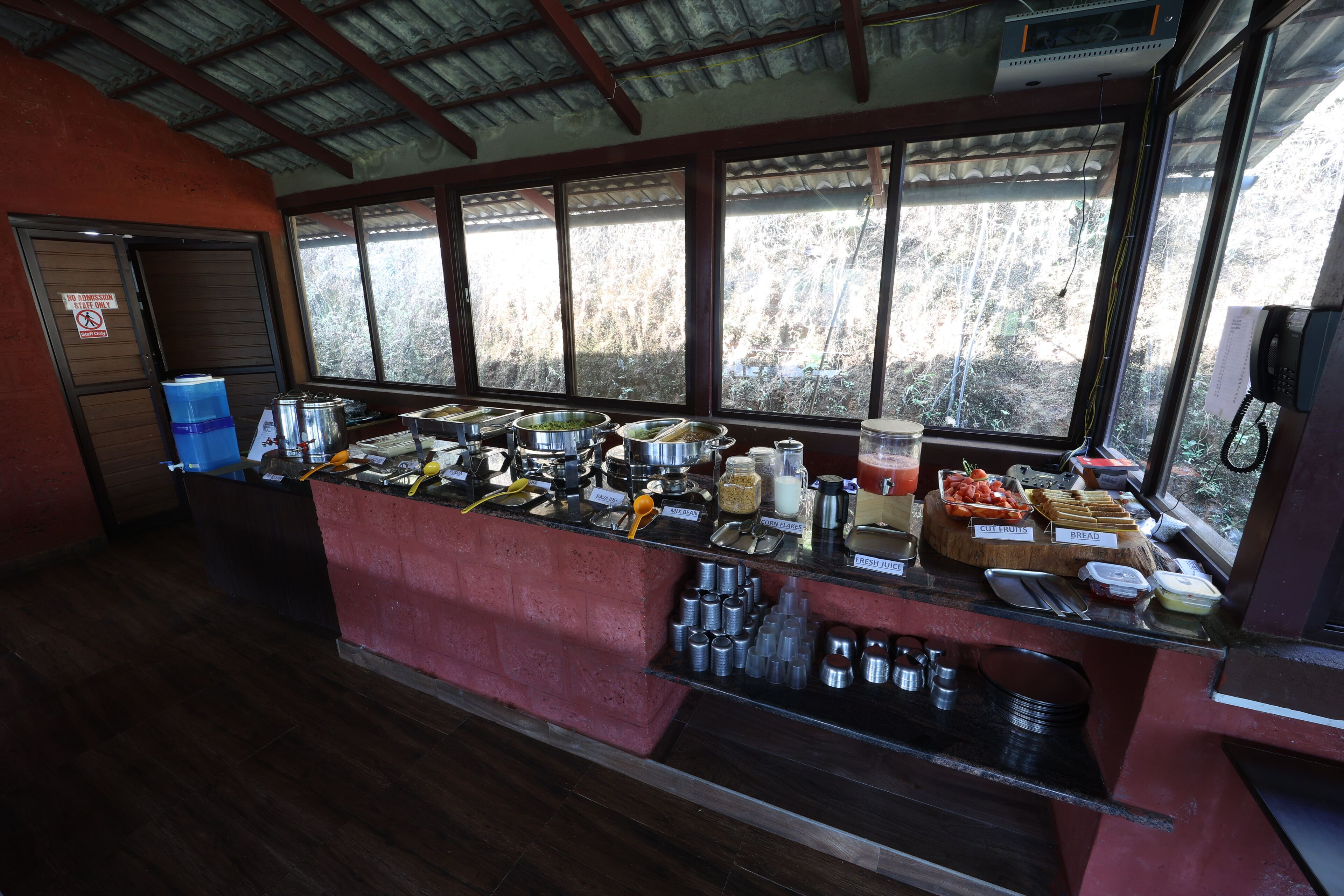Un mostrador de buffet con diversas opciones de comida y bebidas, un teléfono y una ventana con vistas a árboles.