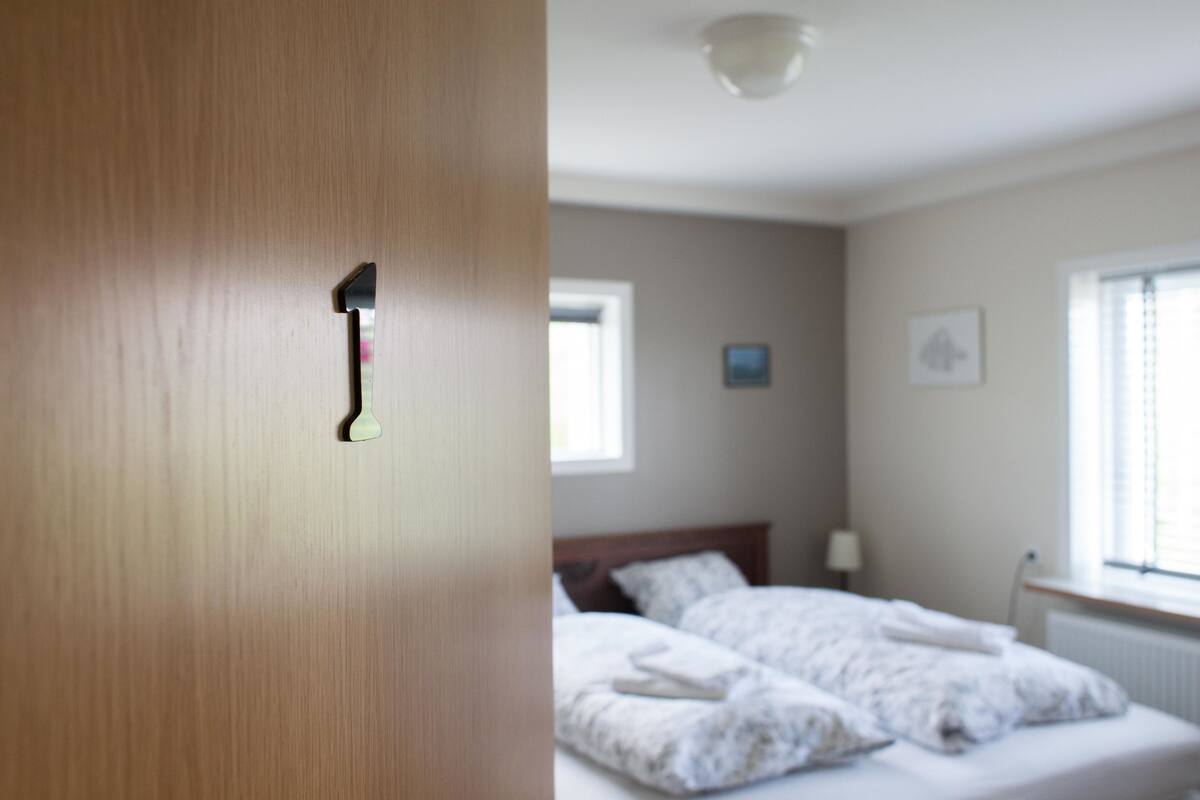 A bedroom with a wooden door, a bed with white bedding, a nightstand, and a window with blinds.