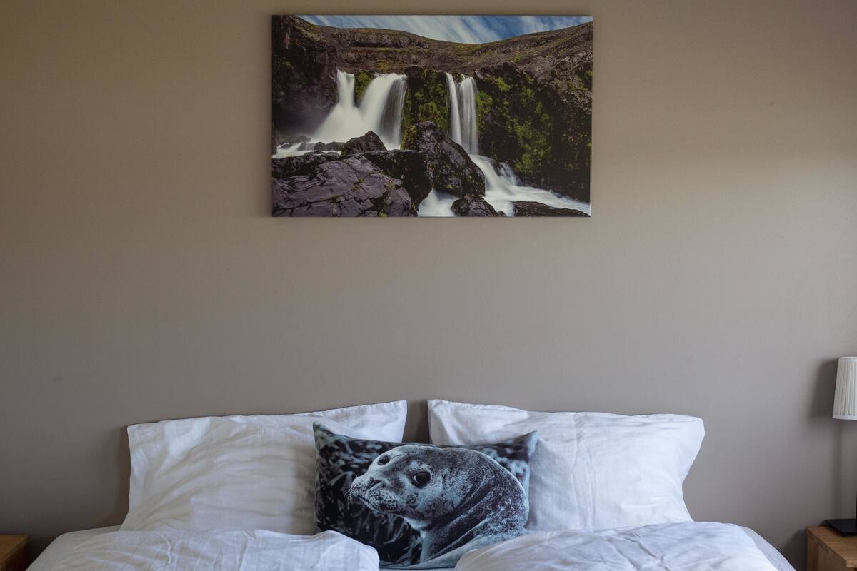 A bedroom with a bed, pillows, a wall-mounted painting of a waterfall, and a bedside lamp.