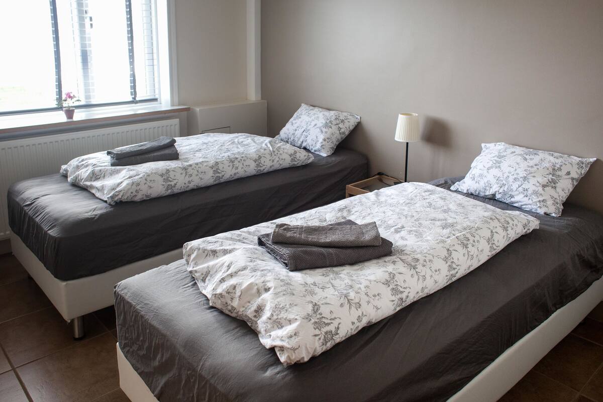 Two single beds with floral bedding, a bedside table with a lamp, and a window with blinds.