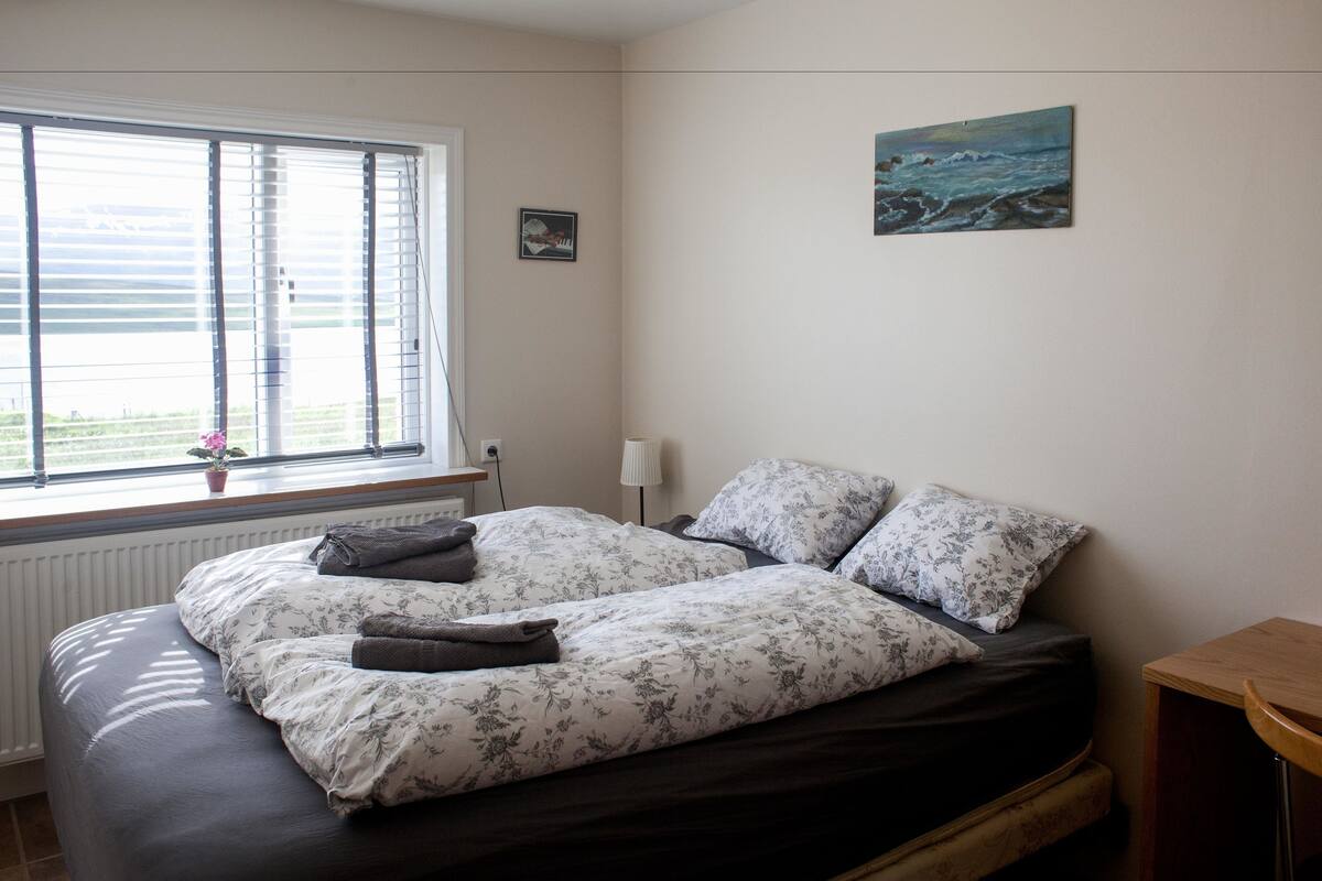 A bedroom with a bed, a window with blinds, a lamp, and a painting on the wall.
