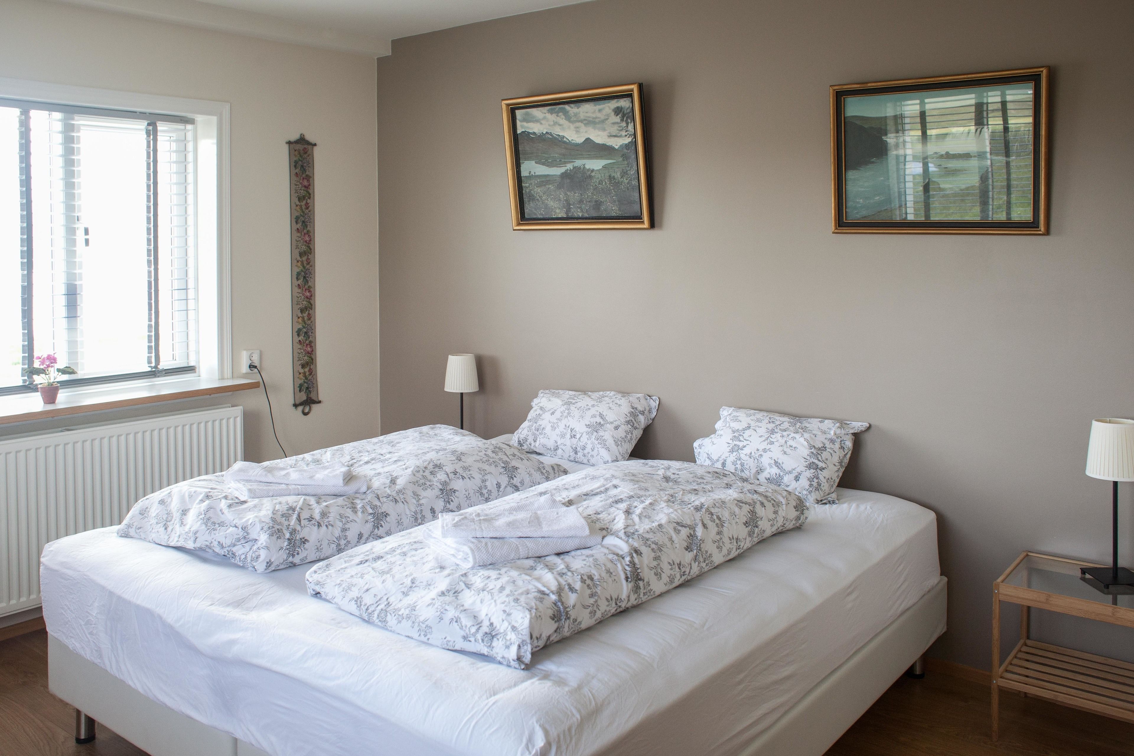 Une chambre à coucher avec un lit double, une literie à motifs floraux, une fenêtre avec des stores, un radiateur, un tableau accroché au mur et une table de chevet avec une lampe.