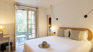 A hotel room with a bed, a desk, a chair, a hat on the bed, a balcony, and a wall-mounted light.