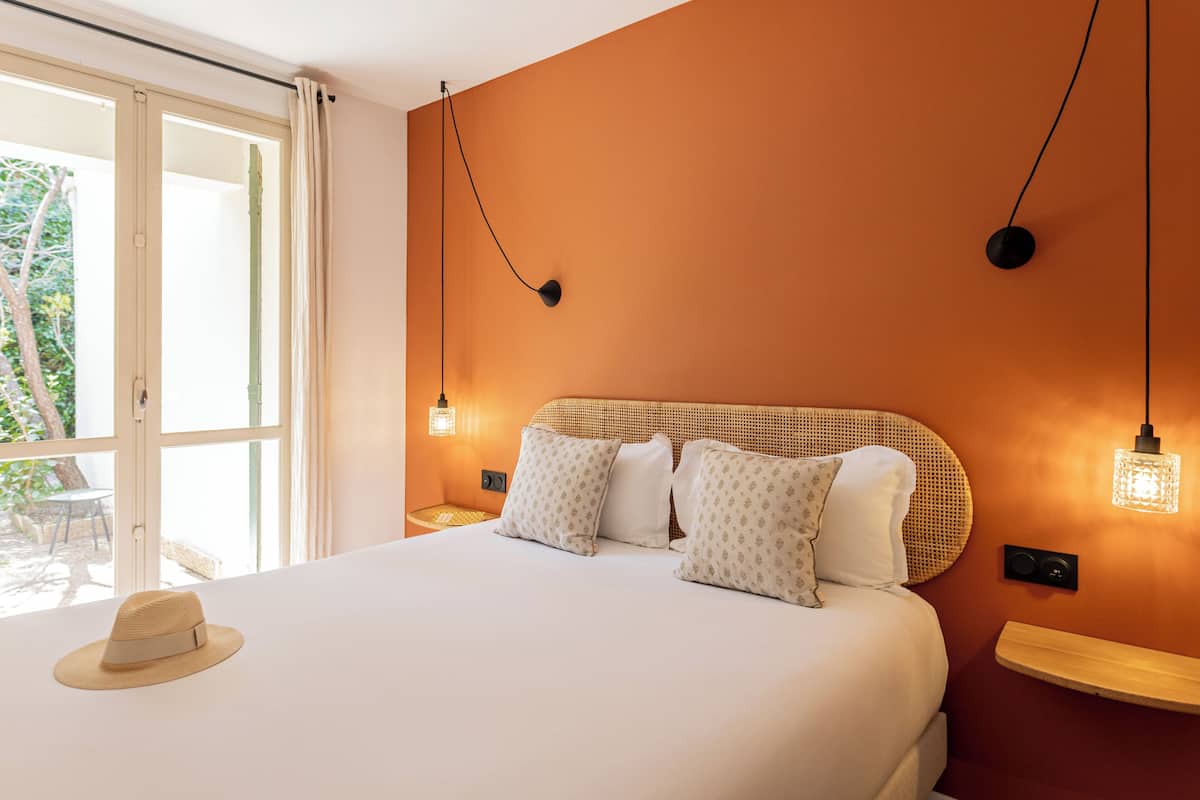 A bedroom with a large bed, a hat on the bed, a window with curtains, and two wall-mounted lights.