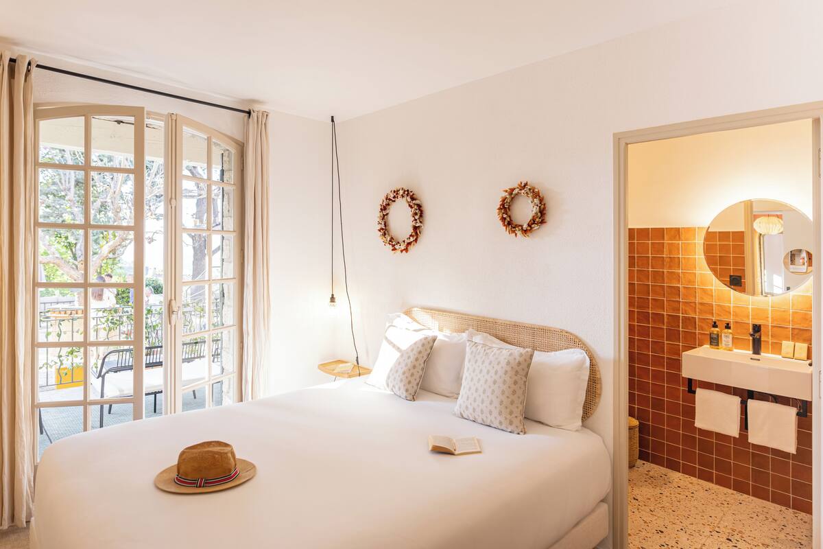 A hotel room with a bed, a hat on the bed, a bathroom with a mirror and sink, and two wreaths on the wall.