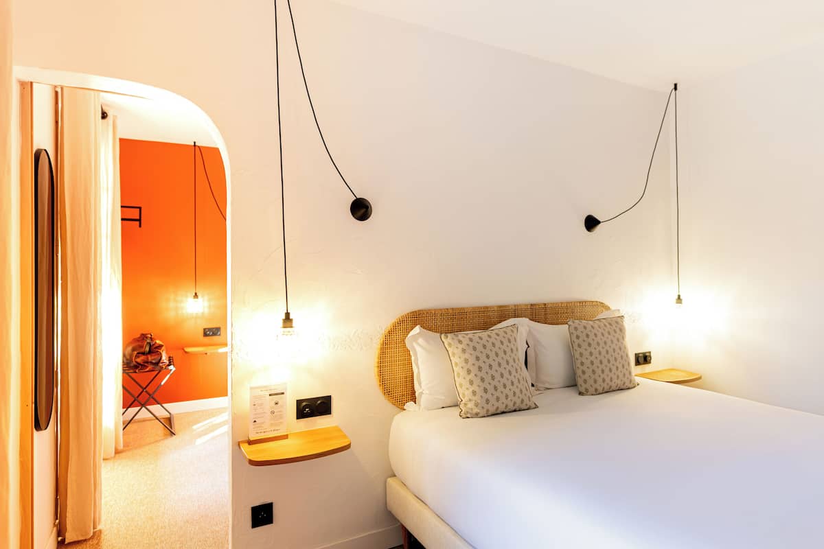 A hotel room with a bed, two bedside tables, a wall-mounted lamp, and a door leading to another room.