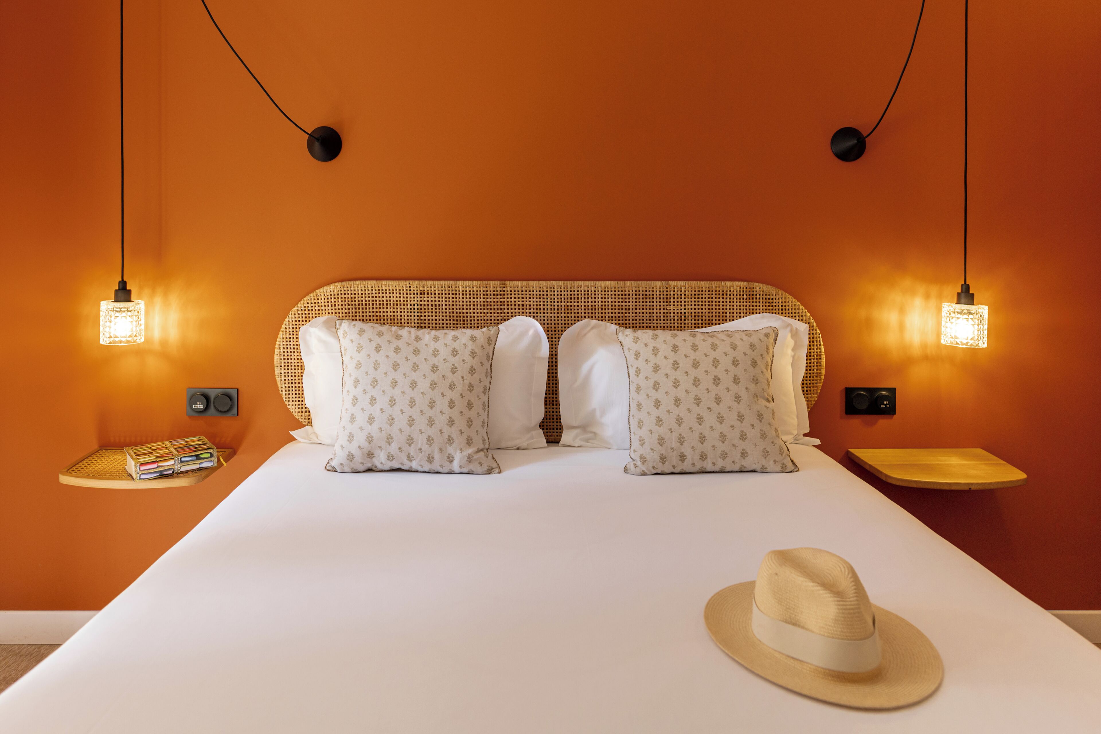 A bed with a woven headboard, two pillows, a bedside table with a book, and a hat on the bed.