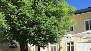 An outdoor patio with wooden flooring, black chairs, and tables. There is a large tree providing shade, and a white fence enclosing the area. - Hotel Ranten (Falkoping)