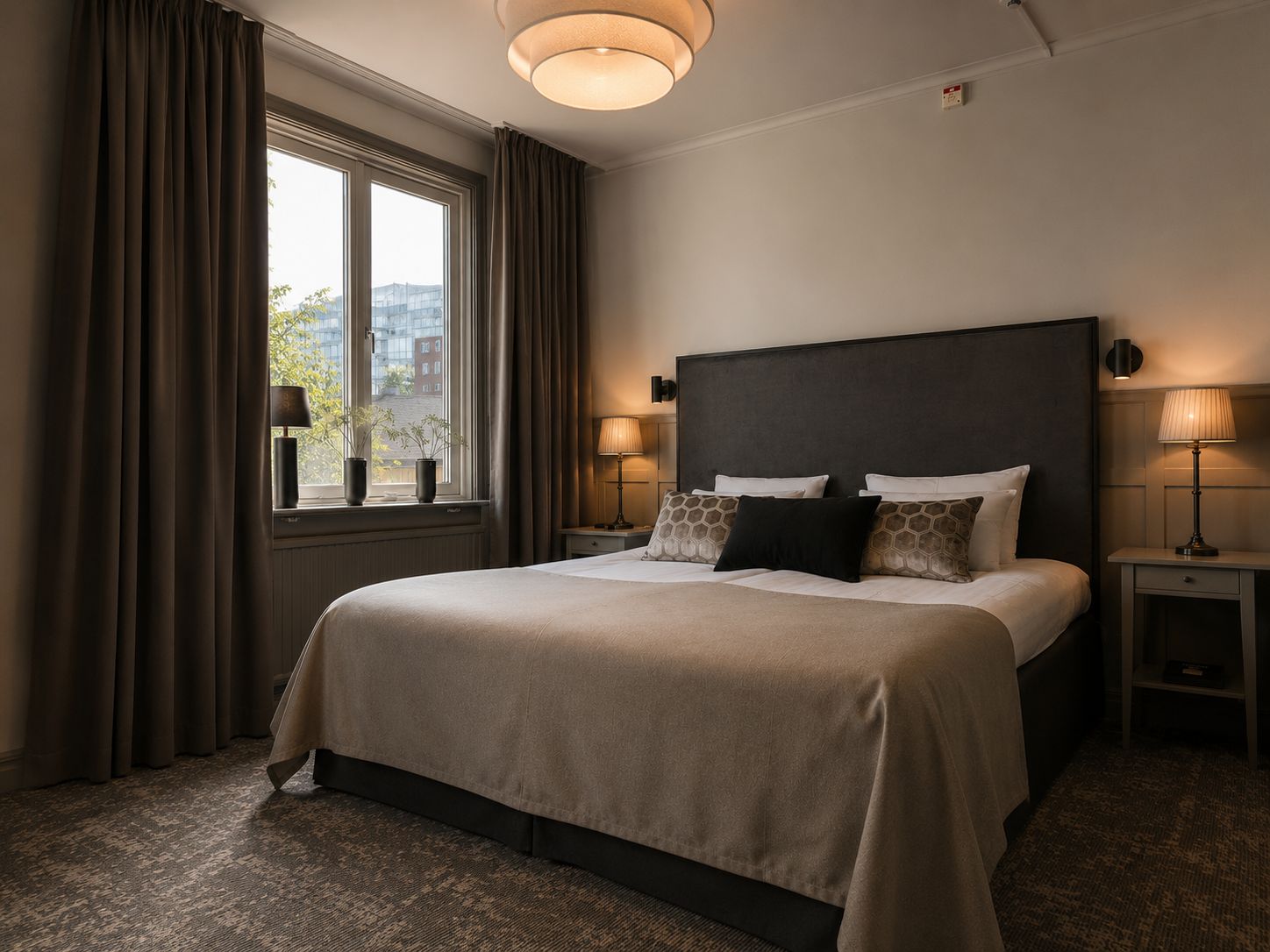 Une chambre d’hôtel avec un grand lit, deux tables de chevet, une fenêtre avec des rideaux et une vue sur les bâtiments environnants.
